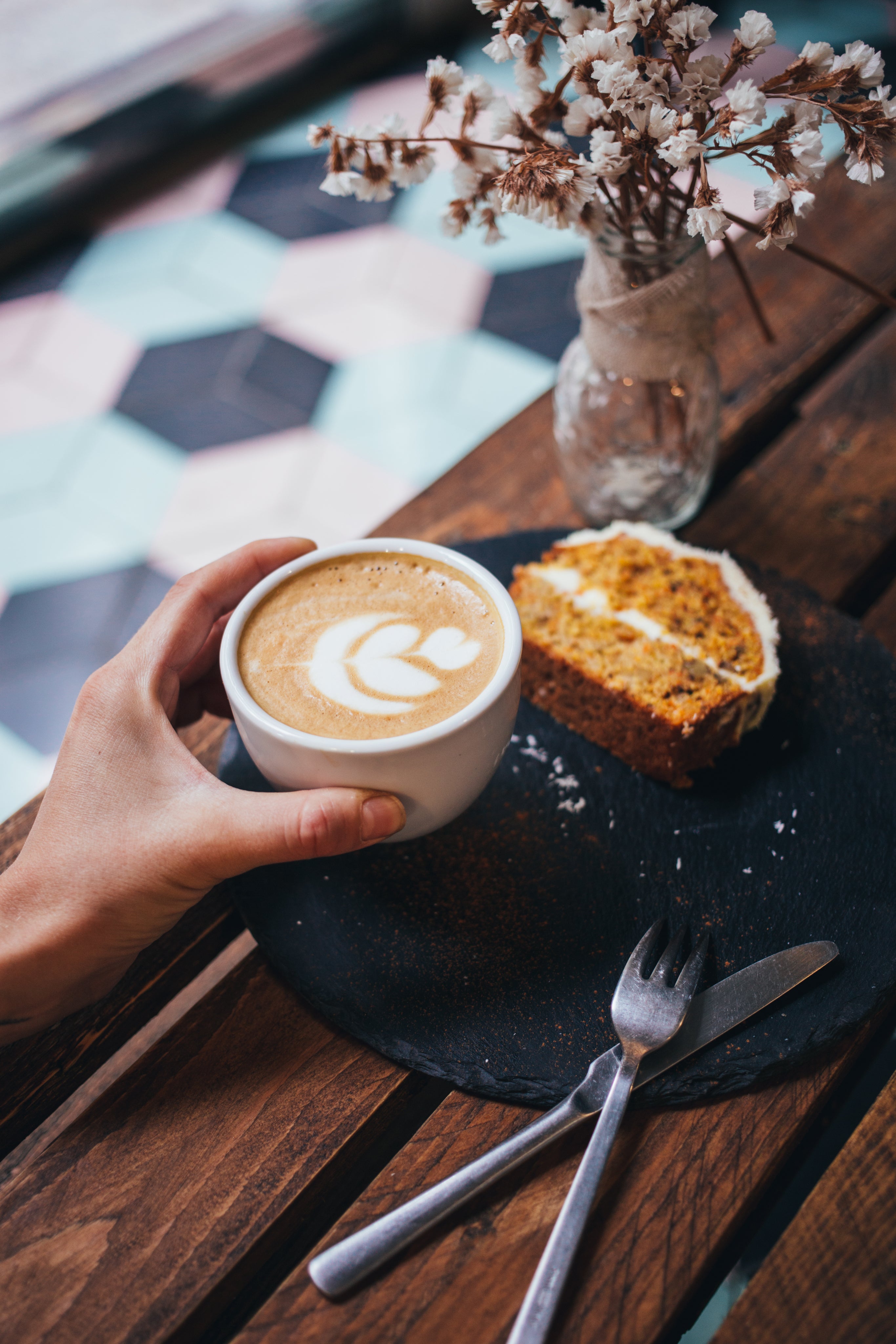 Latte art on a coffee cup with carrot cake slice on a slate plate and utensils on a wooden table